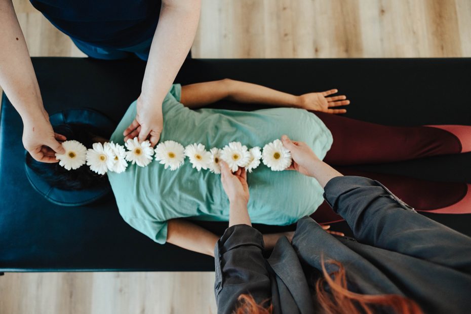 A client lying face down with flowers being laid along their spine by two Embodied staff