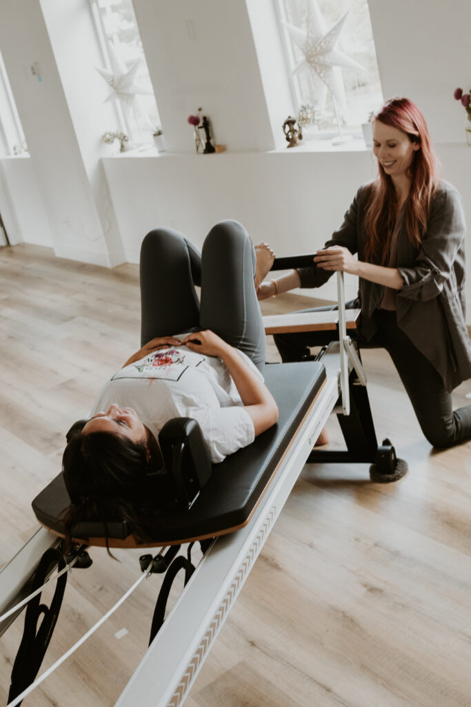 A client lying on the reformer on their back with bent knees and Katie kneeling by their feet