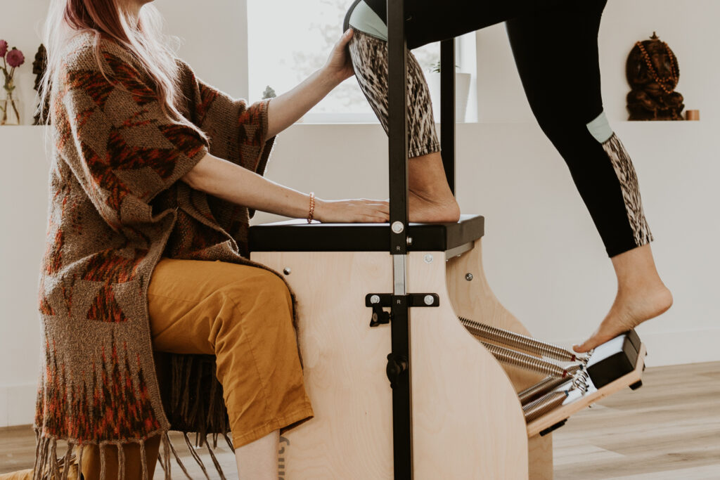 A closeup showing the lower legs of client taking steps on the Pilates Chair, Katie is visible from the neck down in a kneeling position facing the client with a hand on their knee and a hand on their foot