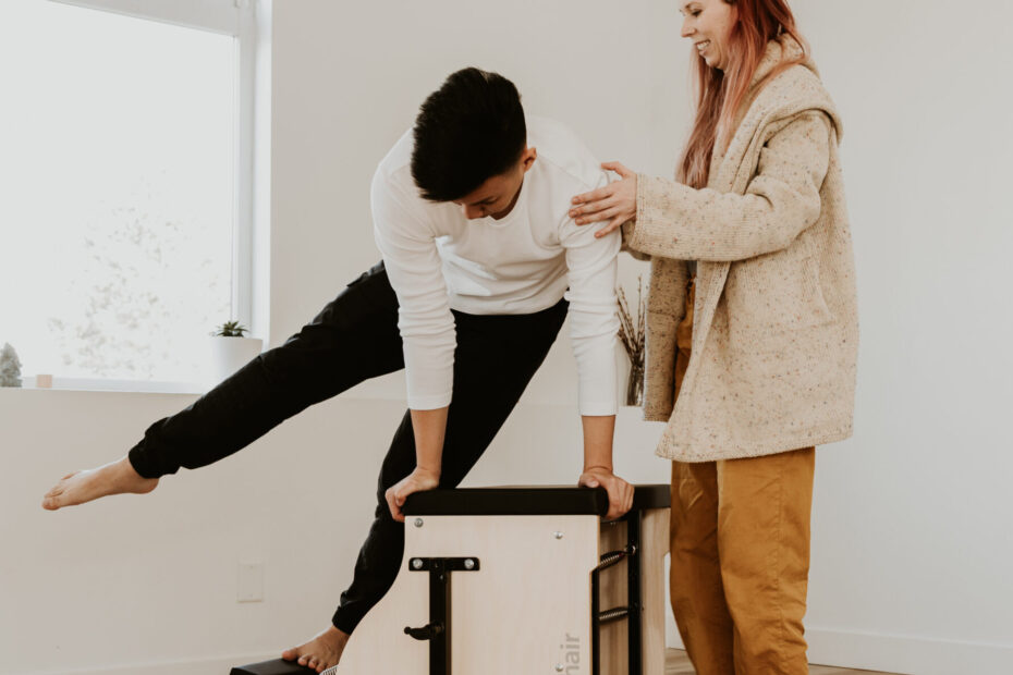 A client with their hands propping them up below their shoulders, their left foot extended to the right also propping them up, and their right leg extended out and lifting beyond that with Katie standing on the client’s left touching their left shoulder