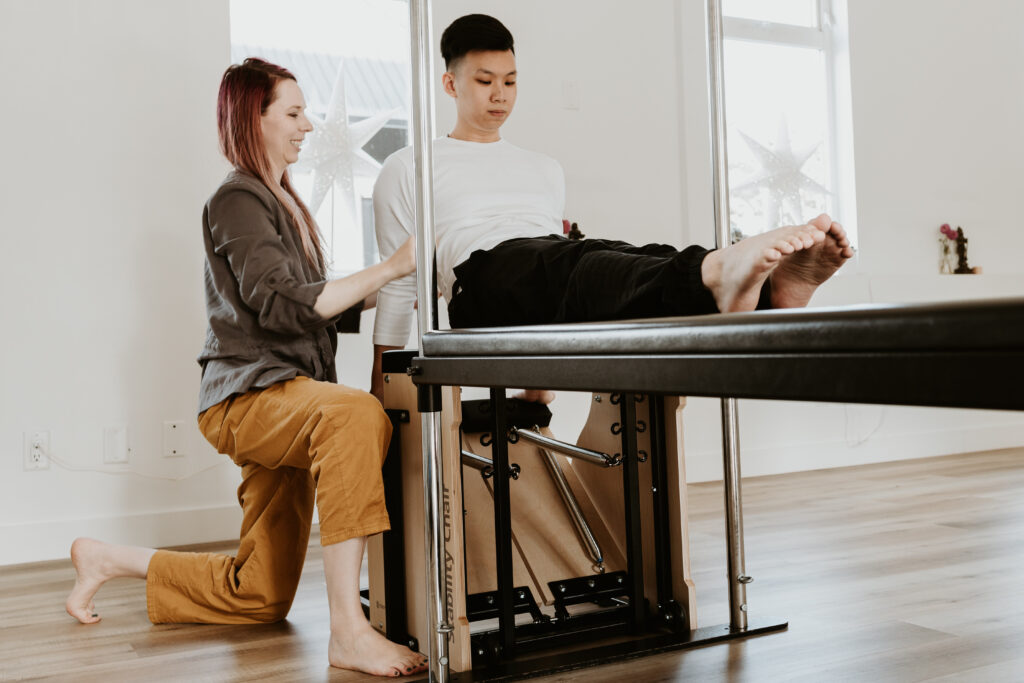A client seated on the Pilates Cadillac with their legs stretched out in front of them and their hands propping them up with Katie kneeling beside them with her hand on their arm