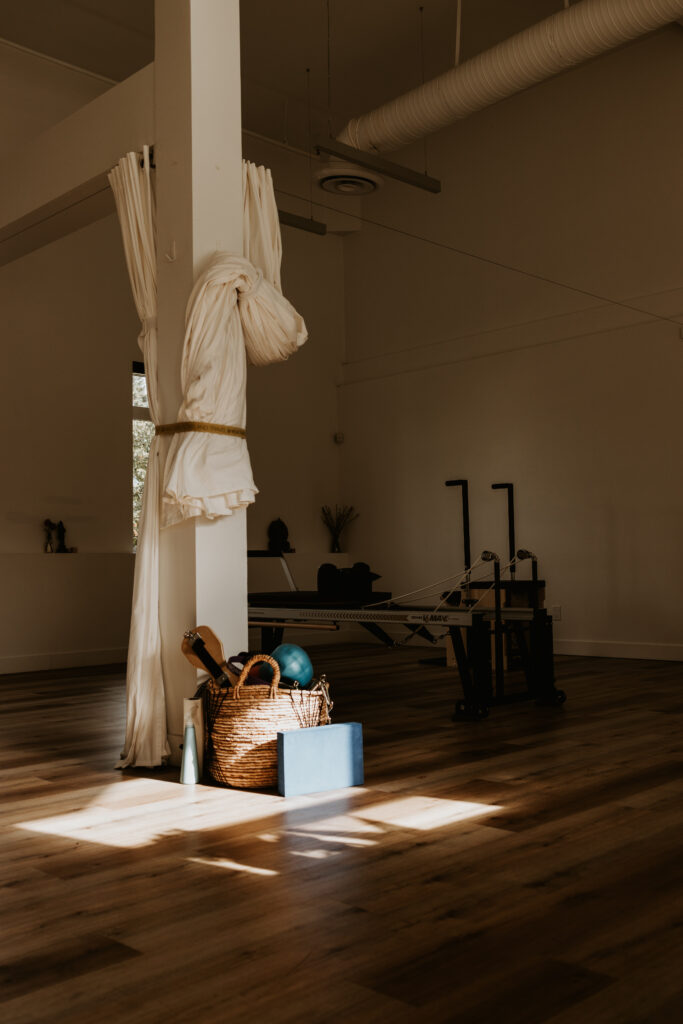 A shot of the studio, in the forefront a pillar with tied up curtains on it, a basket of Pilates equipment at the base of the column, and light shining on the basket and floor with the rest of the shot in moody shadows