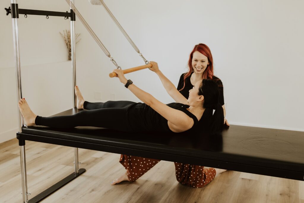 Katie smiling next to a smiling client who is lying on their back on the Pilates Cadillac with their arms outstretched and holding a roll down bar