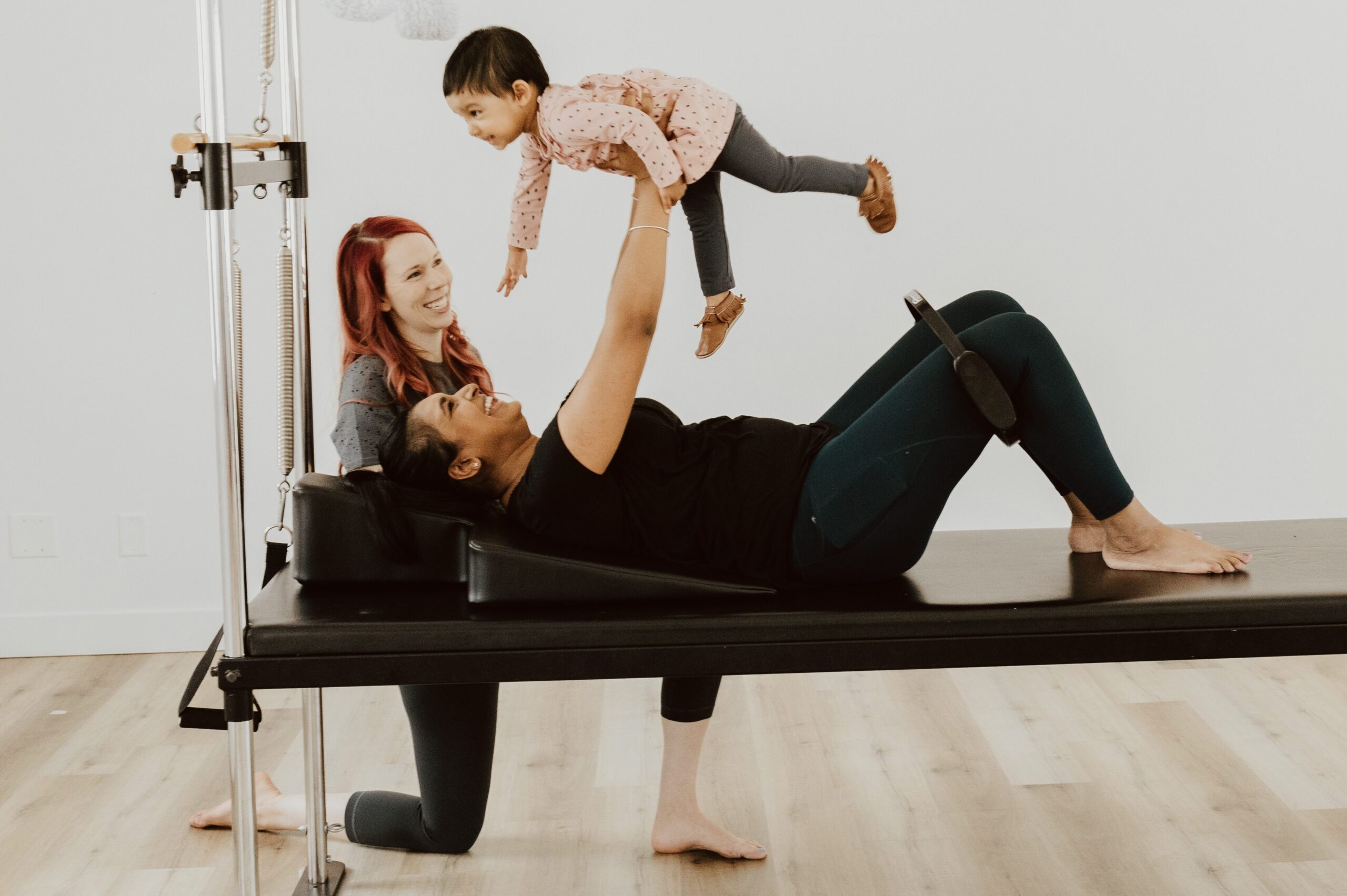 A client lying on their back on the pilates cadillac holding their toddler in extended arms above them with Katie kneeling beside them