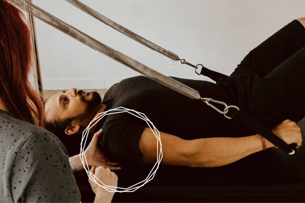 A closeup of a client lying on their back on the Cadillac. Katie is correcting the position of the right shoulder, while client is pulling down on the springs, with their hands at their sides