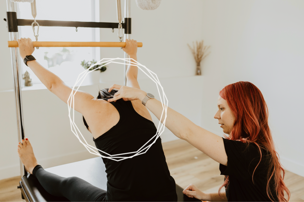 Katie working with a client who is in a seated forward fold on the Pilates Cadillac with their hands outstretched holding a roll down bar. Katie making an adjustment with one hand on the client's upper back