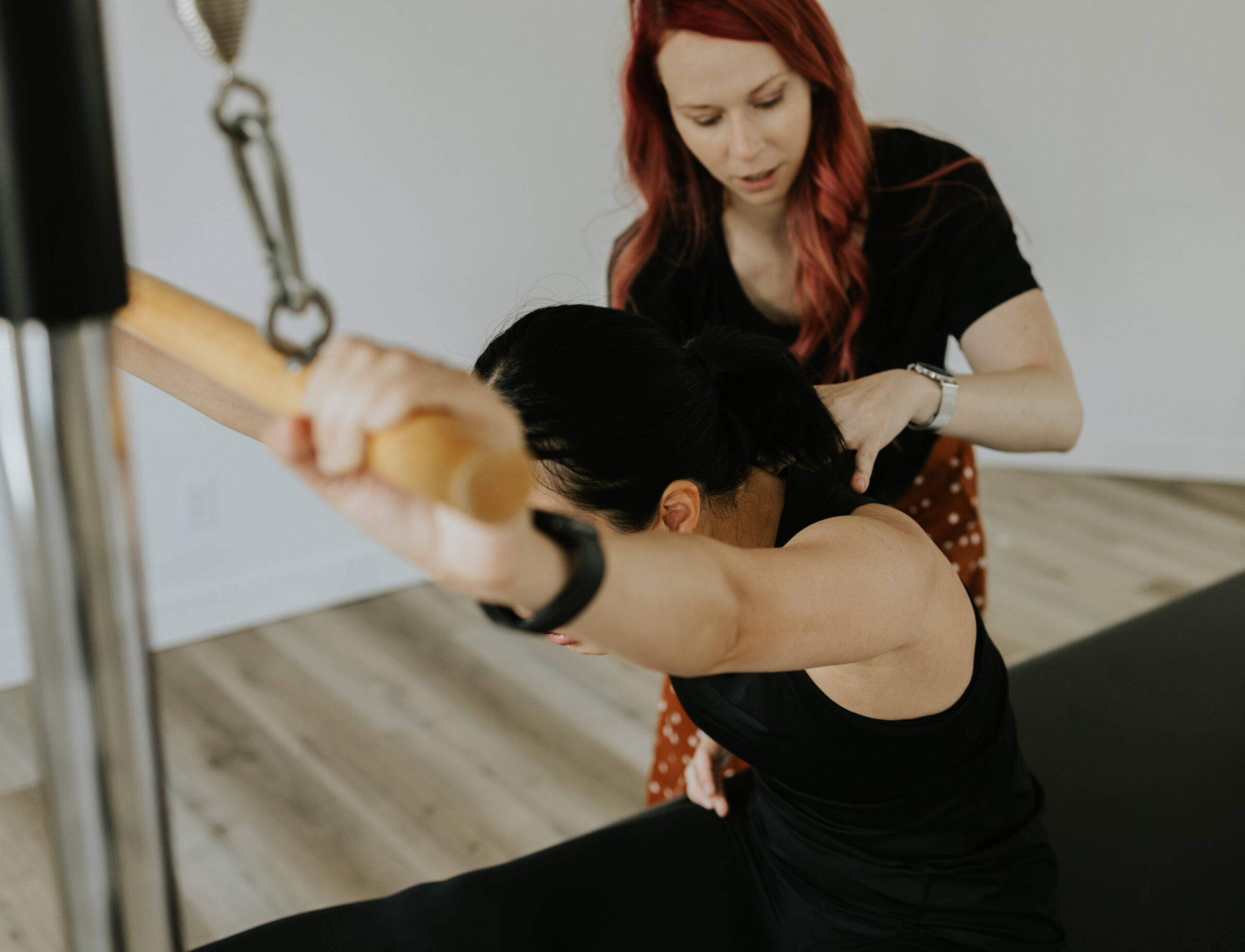 A physiotherapist working with a client who is in a seated forward fold on a Pilates Cadillac with hands holding a bar diagonally forward above their head